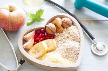 heart-shaped bowl filled with healthy foods and surrounded by a stethoscope, an apple, and a dumbell